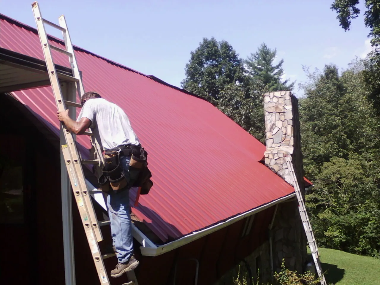 Roofer on a ladder beside a red metal roof and stone chimney — Soffit & Fascia Repair by Roofing Brothers in South Solon